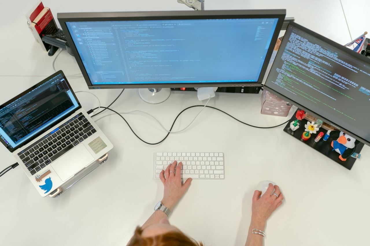Overhead view of a person coding on dual monitors and a laptop at a white desk.
